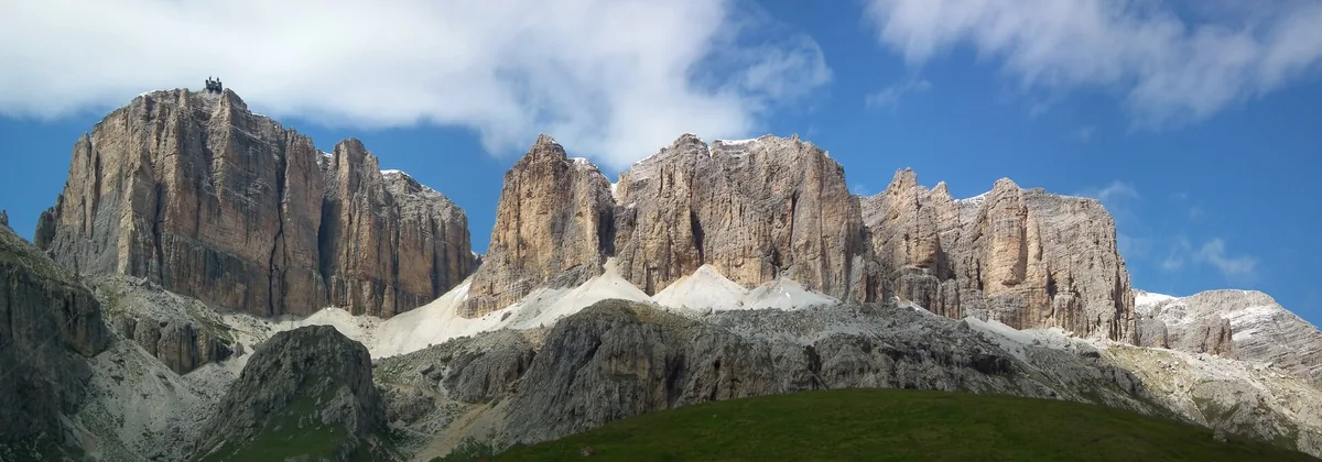 Grey Dolomite cliffs rising above a green grass slope under a blue sky