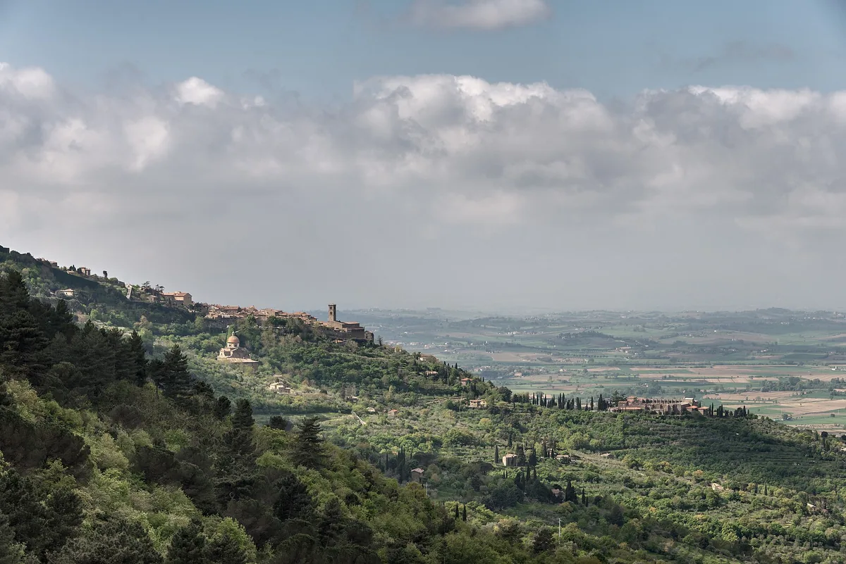 A hilltop town on a wooded ridge above a broad farming valley under white clouds