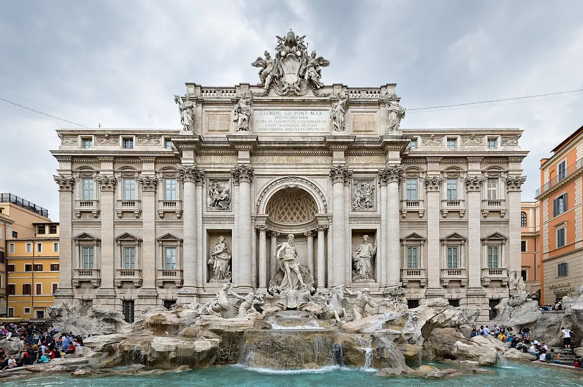 A baroque marble fountain with statues and a turquoise pool below a palace facade