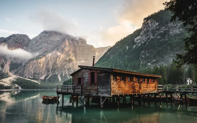 A wooden boathouse on green water below a rock face and pine forest