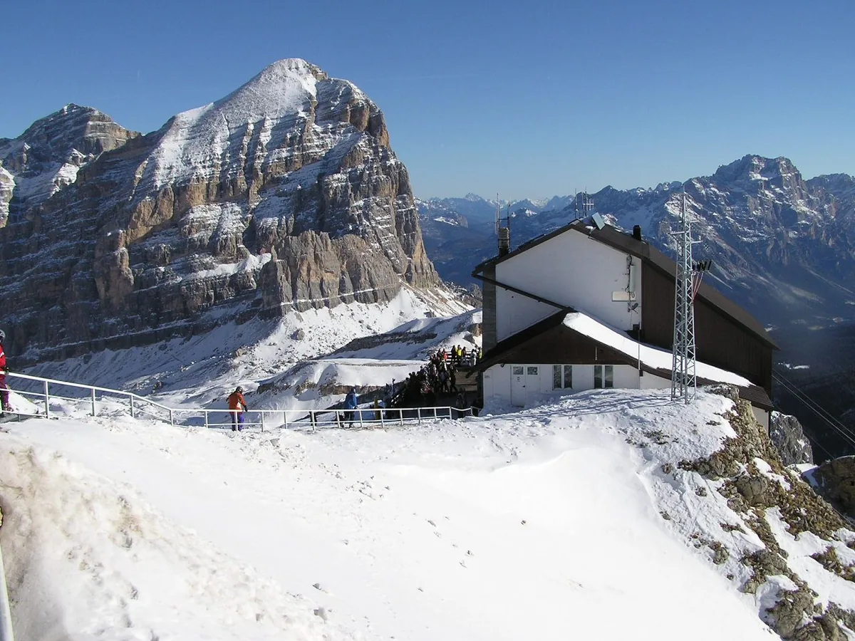 A mountain hut with a sloping roof on a snowy ridge facing Dolomite peaks