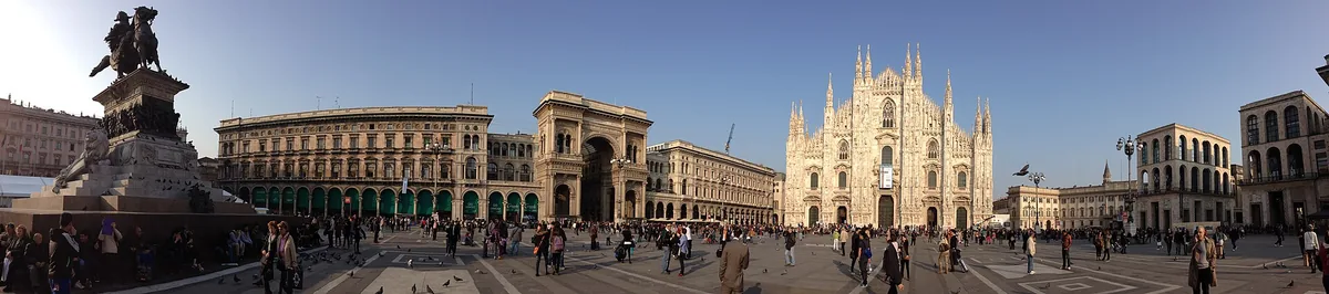 Wide paved square with an arched gateway, strolling crowds and a white Gothic cathedral to the side