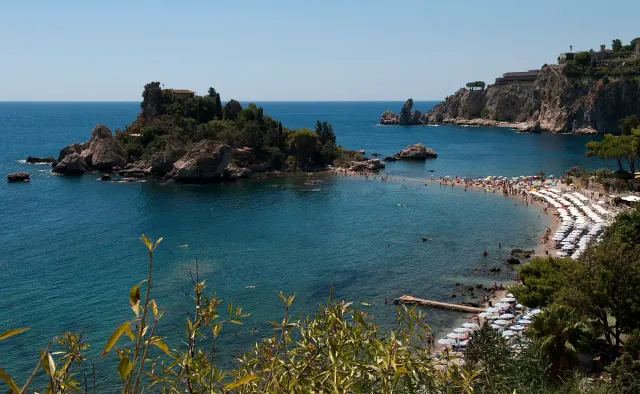 A green rocky islet in turquoise water beside a pebble beach
