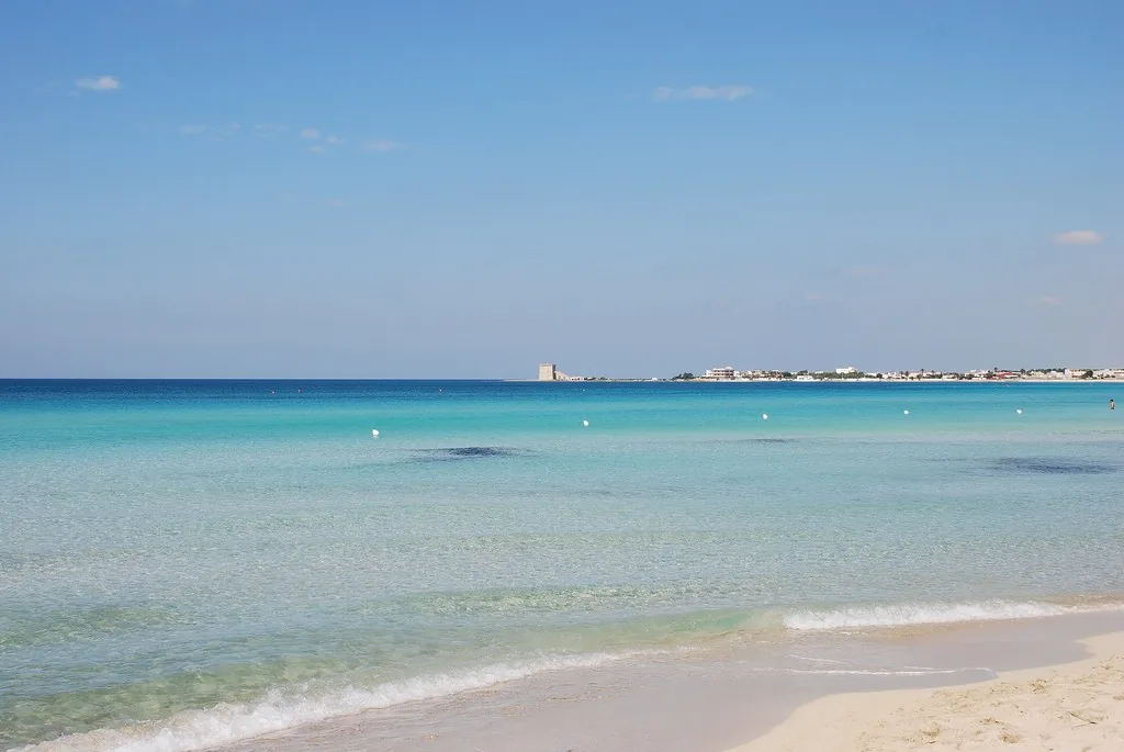 A pale sand beach with shallow turquoise water and a small watchtower on the horizon