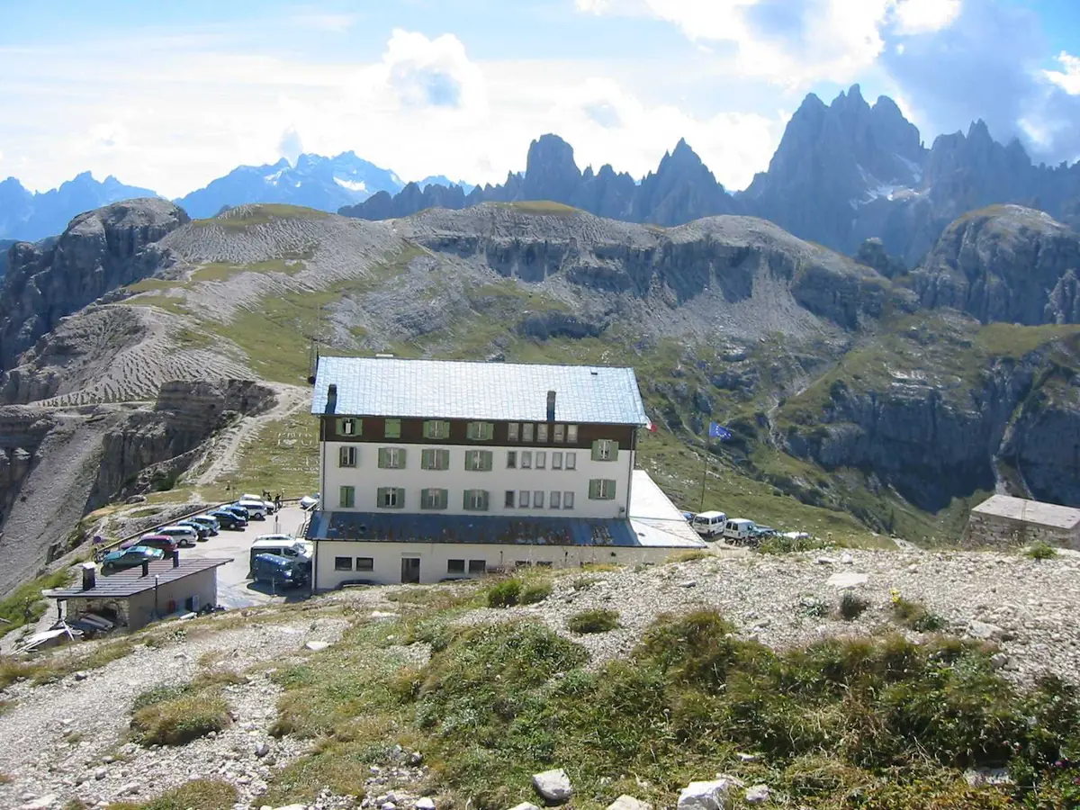 A long stone building with a pale roof on a mountain saddle, jagged peaks behind