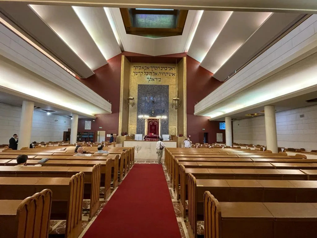 A prayer hall with wooden benches in two rows, a red carpet aisle and an ark at the end