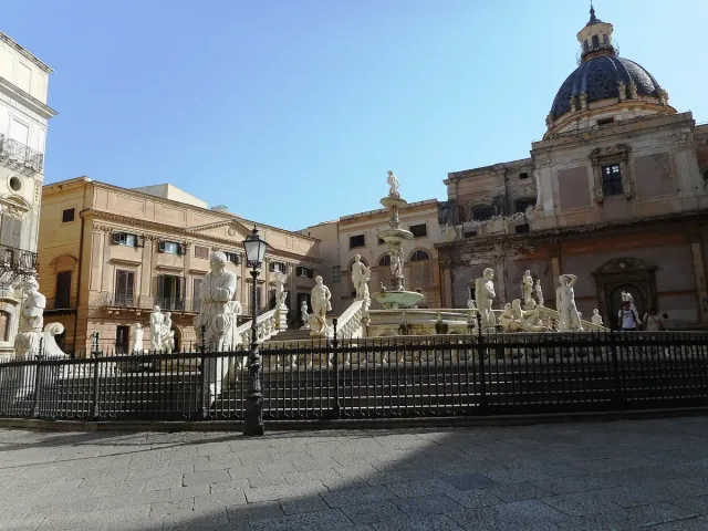 A round fountain of white marble statues and steps in a city square