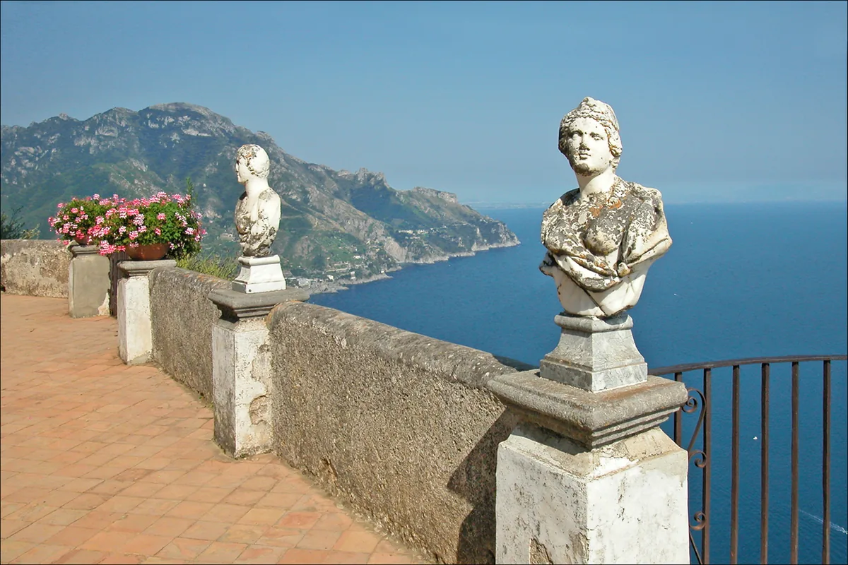 A terrace balustrade with marble busts on plinths above a blue bay and green coastal cliffs
