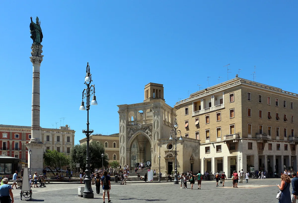 A paved square under blue sky with a tall statue topped column and an arched stone building