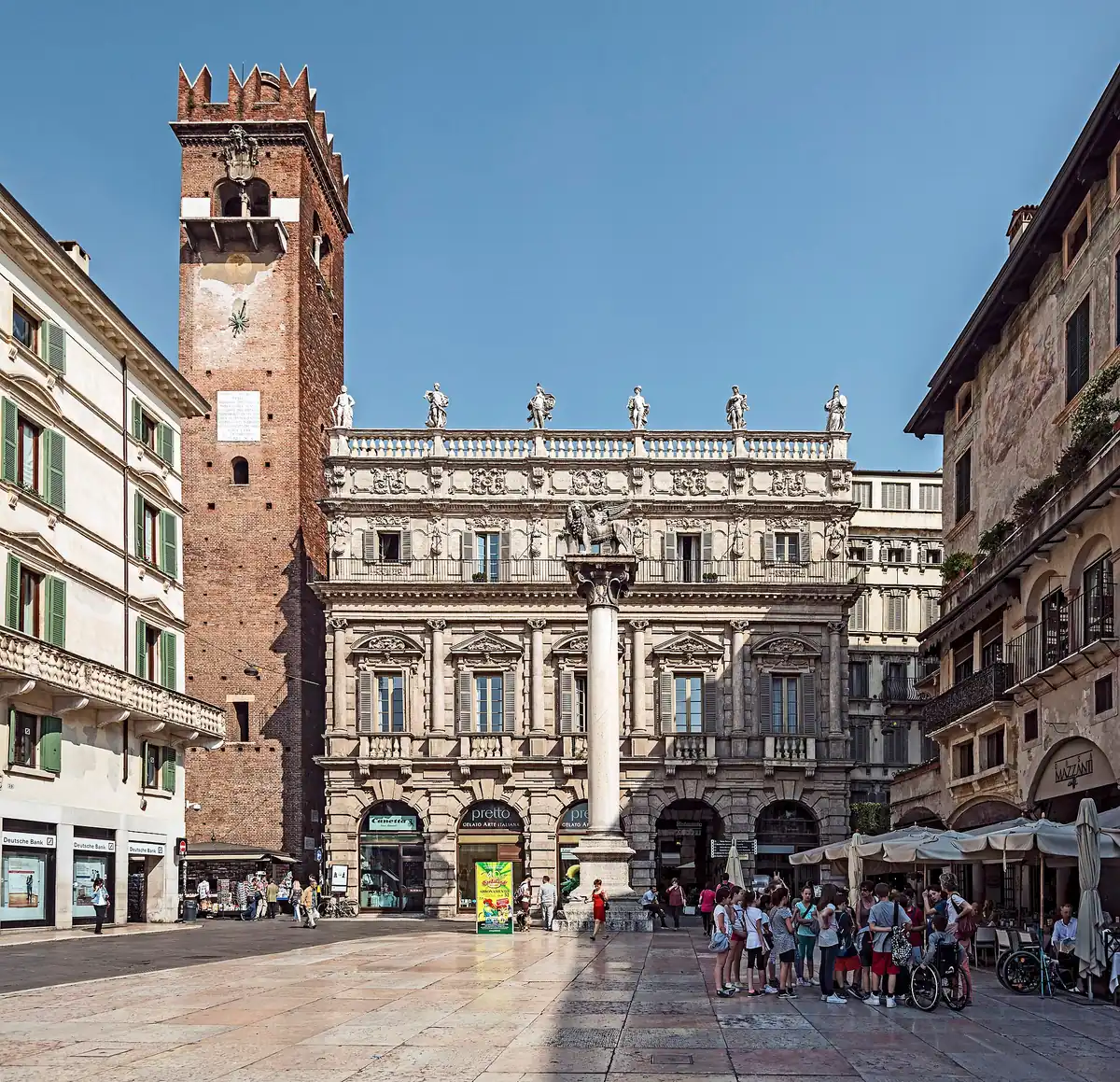 A square with a marble column carrying a statue, a decorated palace and a tall brick tower
