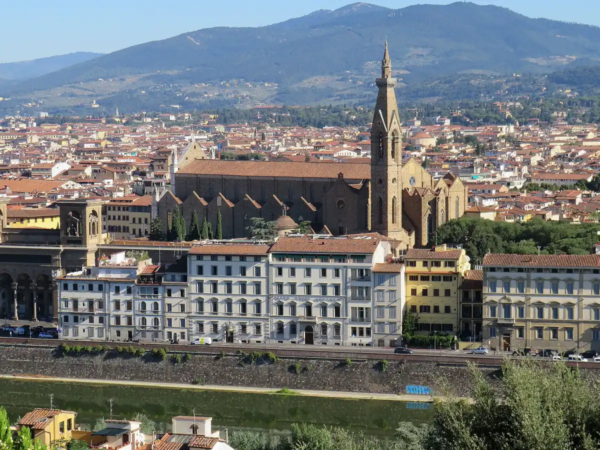 A Gothic basilica with a pointed bell tower above tiled roofs and a river bank