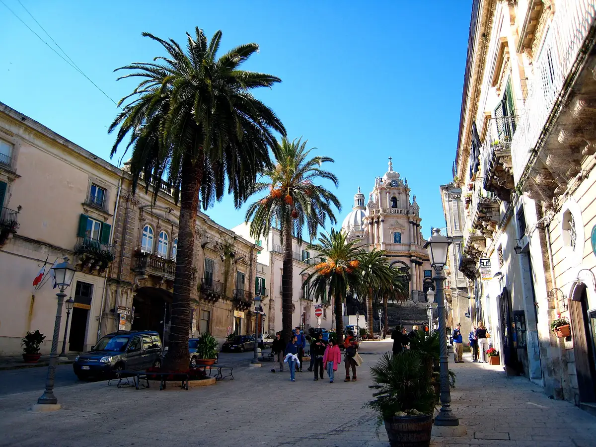 A square with tall palms and a baroque church front with a bell tower at its end
