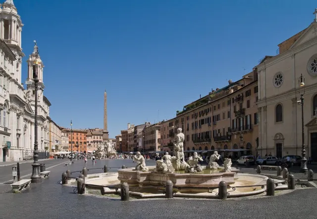 An elongated square with a marble fountain and obelisk ringed by sand coloured houses