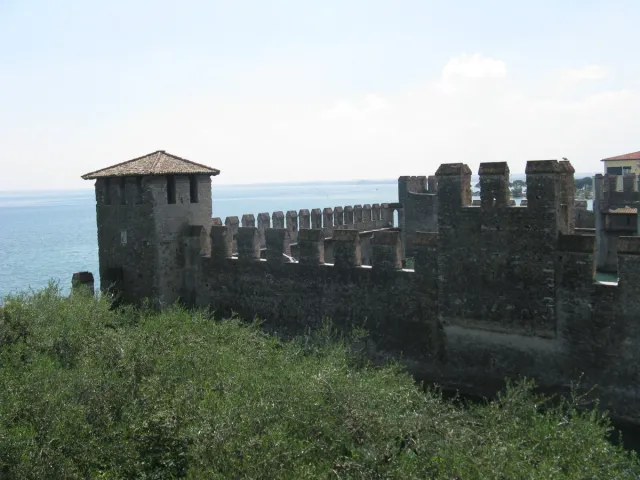 Grey battlemented castle walls above scrub and blue water
