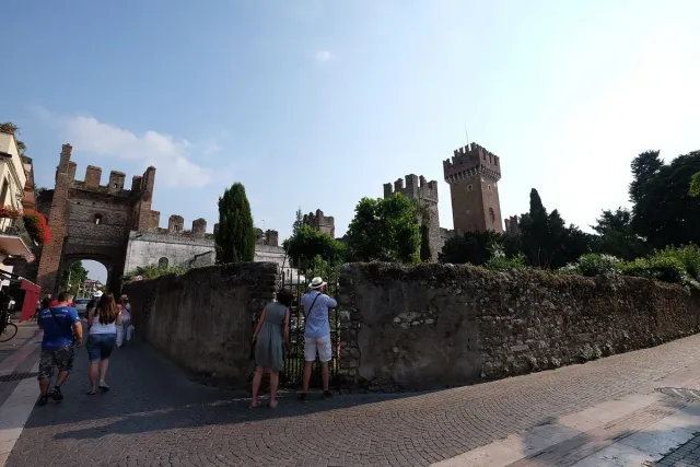 A battlemented town wall beside a lakeside promenade with strollers and trees