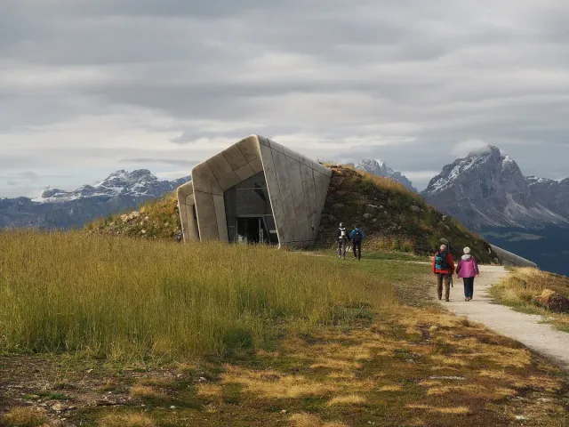 A concrete entrance set into a mountain summit with hikers and snowy peaks behind