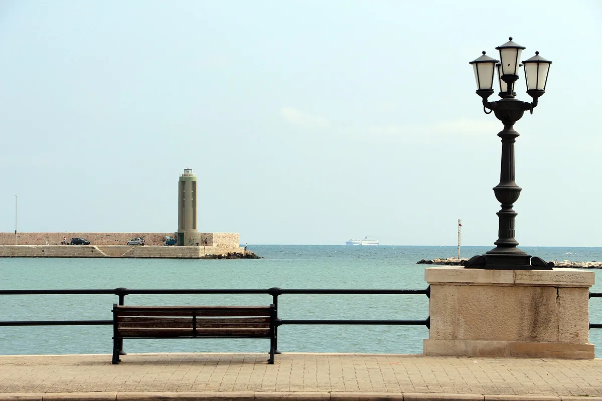 A seafront promenade with a bench and an ornate iron lamp, a breakwater and lighthouse behind