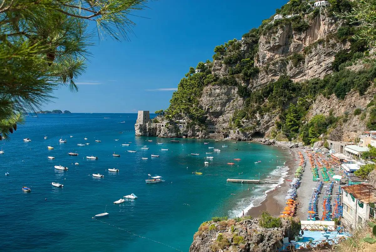Turquoise bay with moored boats, rows of parasols on the sand and a wooded cliff above