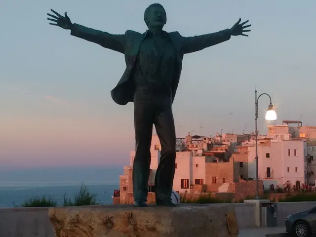 A bronze statue of a man with open arms facing a coastal town and the sea