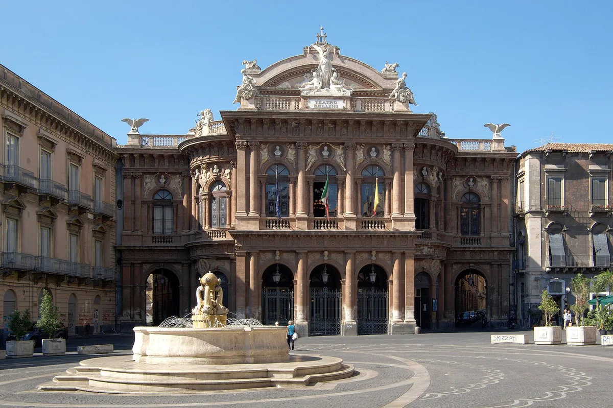 Baroque theatre facade in rosy stone with a stone fountain on the square in front