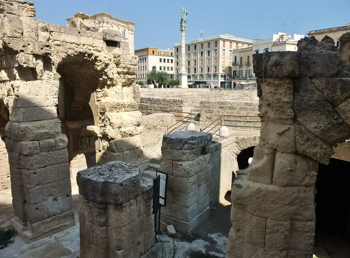 Excavated Roman amphitheatre with tiered seating and column blocks, modern buildings behind
