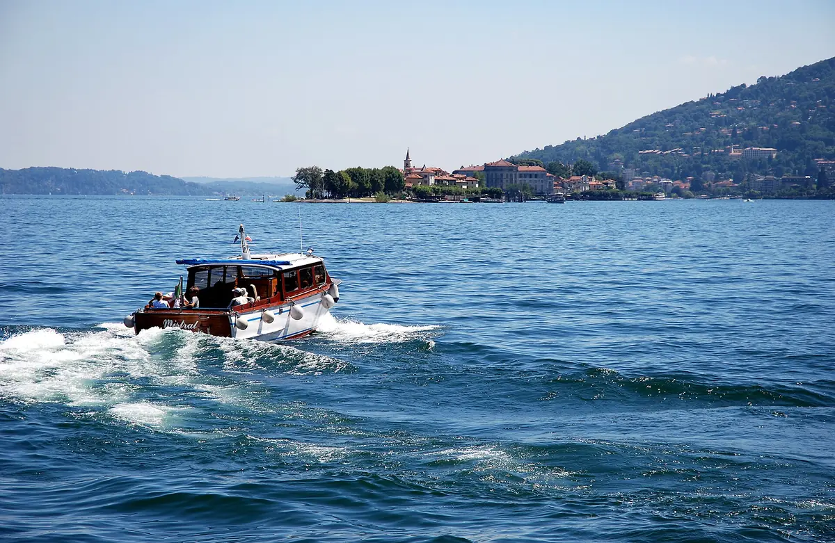 A wooden boat crossing a blue lake with a small island of houses and a tower and green hills behind