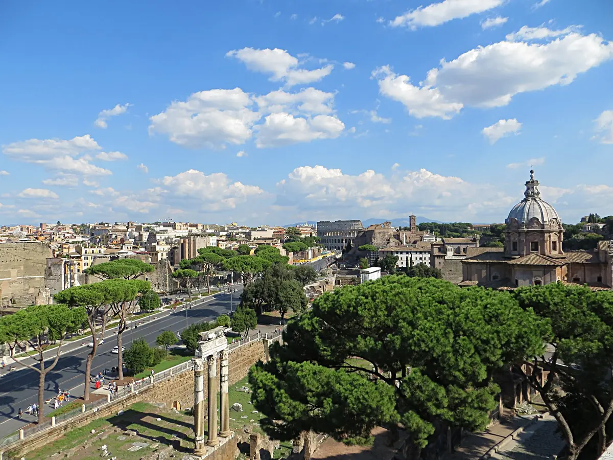 An elevated view over an avenue between Roman ruins, pine trees and a church dome under blue sky