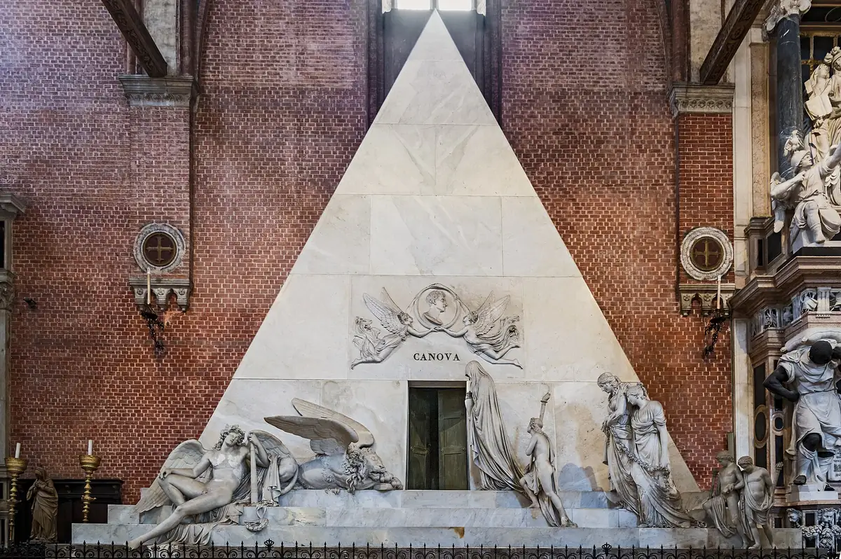 A white marble pyramid tomb with carved figures against a brick wall inside a nave