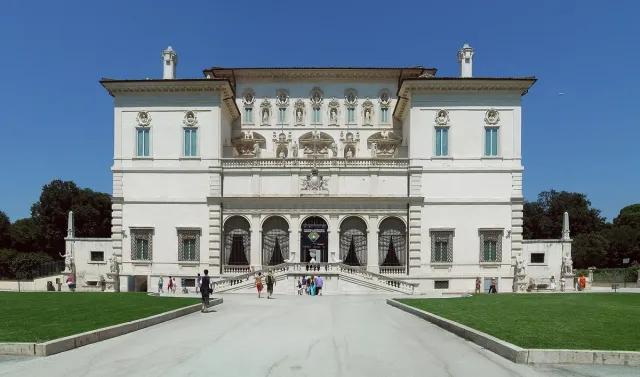A symmetrical white palace with columns and statues above a drive and lawns