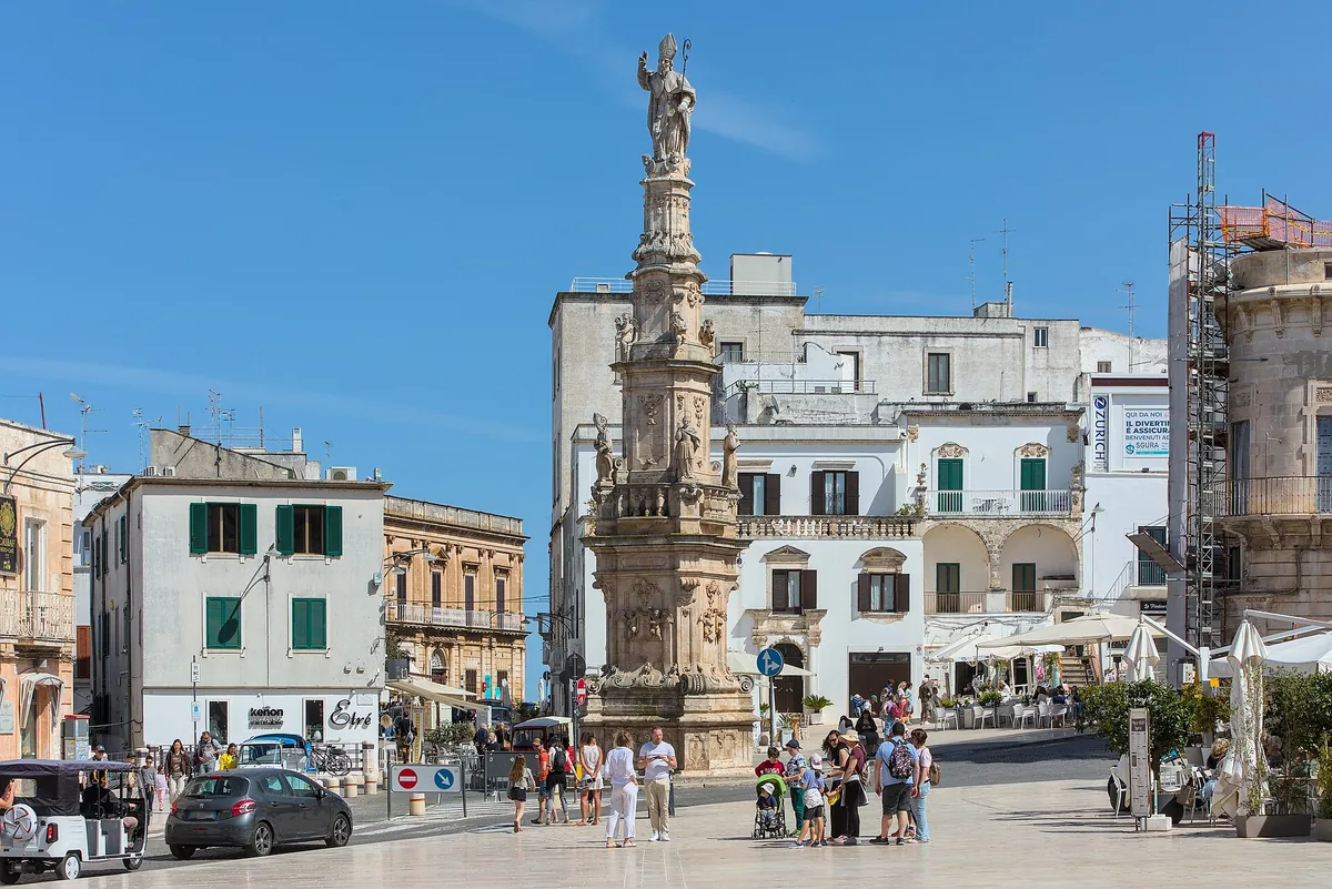 A carved stone obelisk at the centre of a square ringed by white houses under a blue sky