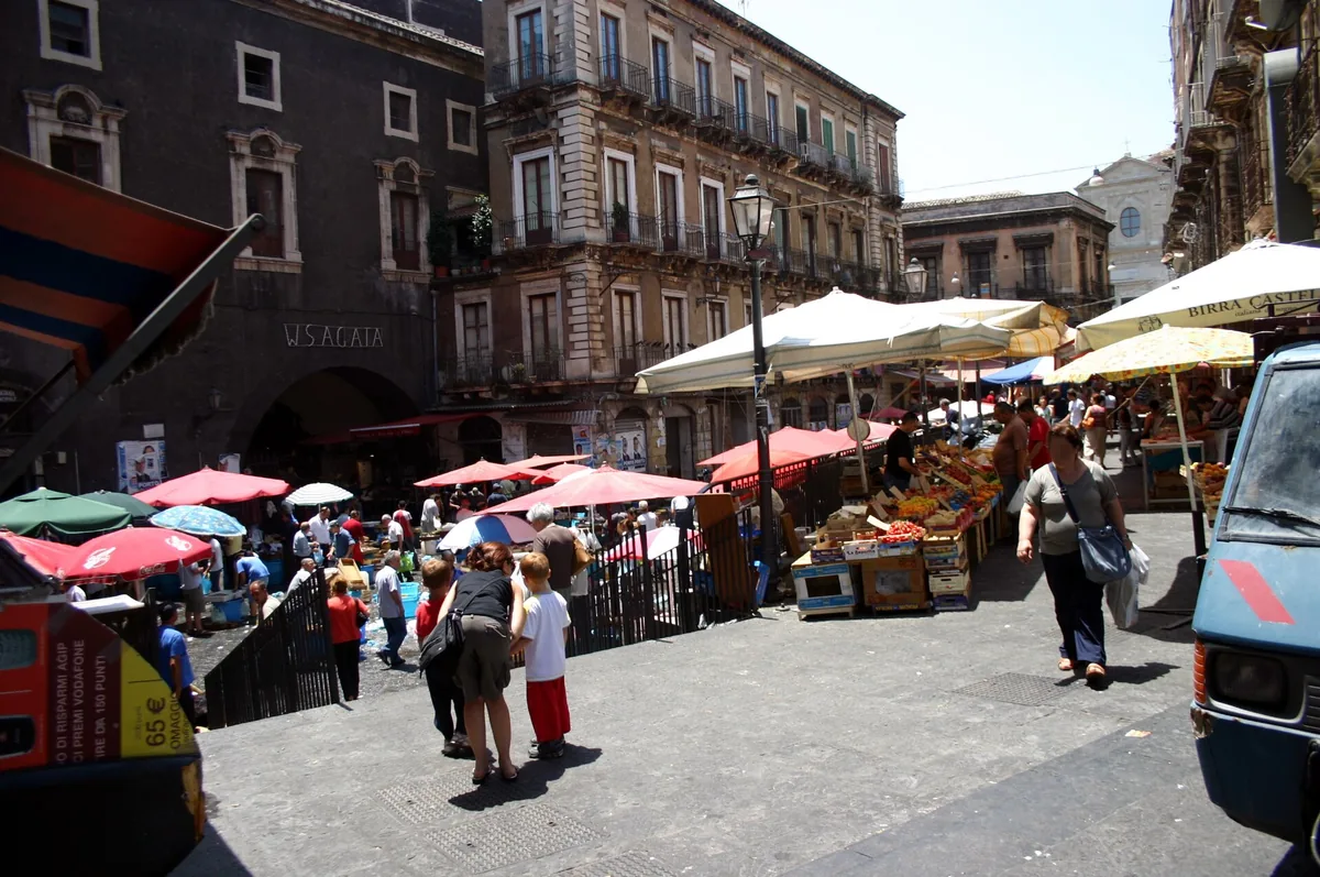 Market stalls under parasols on a paved square before stone facades, with shoppers between them