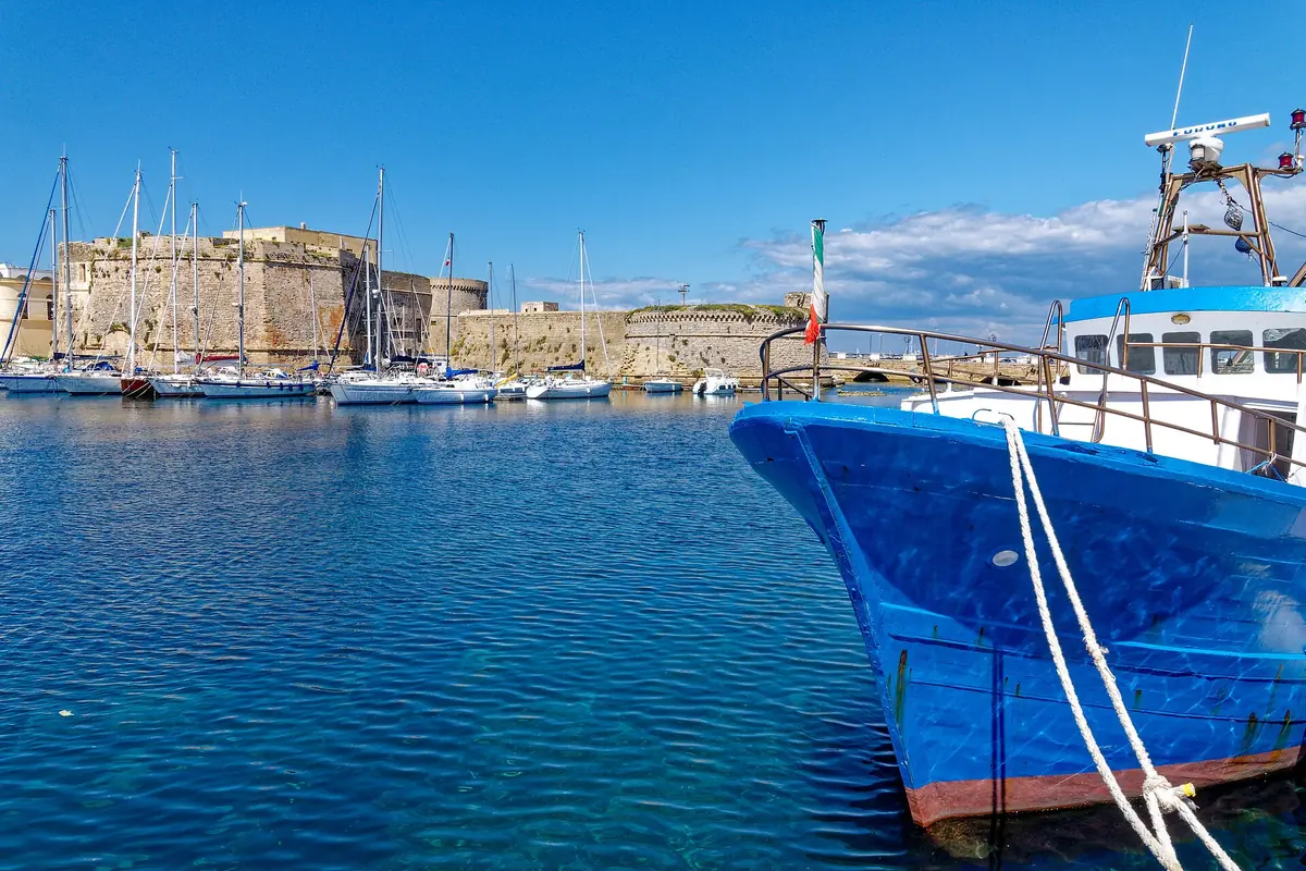 An old town wall above turquoise water, with a blue fishing boat moored in the harbour