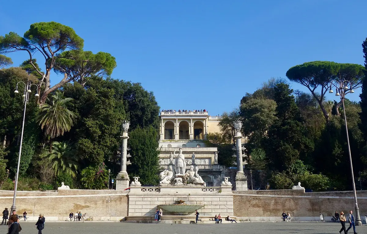 A terrace with a stone balustrade and carved fountain below umbrella pines and a yellow loggia building