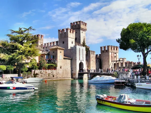 A battlemented castle above a boat filled channel and trees